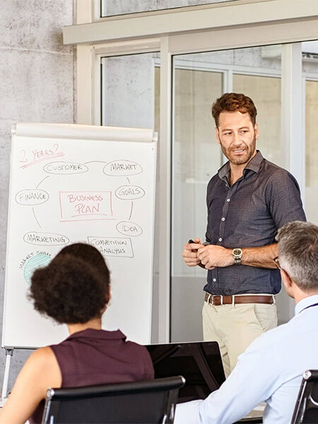 Auf dem Bild sieht man einen Mann der neben einem Flipchart steht. Vor ihm sitzen zwei Personen, die ihm zuhören.