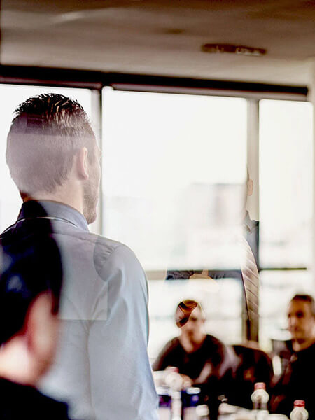 Auf dem Bild sieht man einen Mann der in einem Konferenzraum vor einer Gruppe Personen steht, die an einem Tisch sitzen.