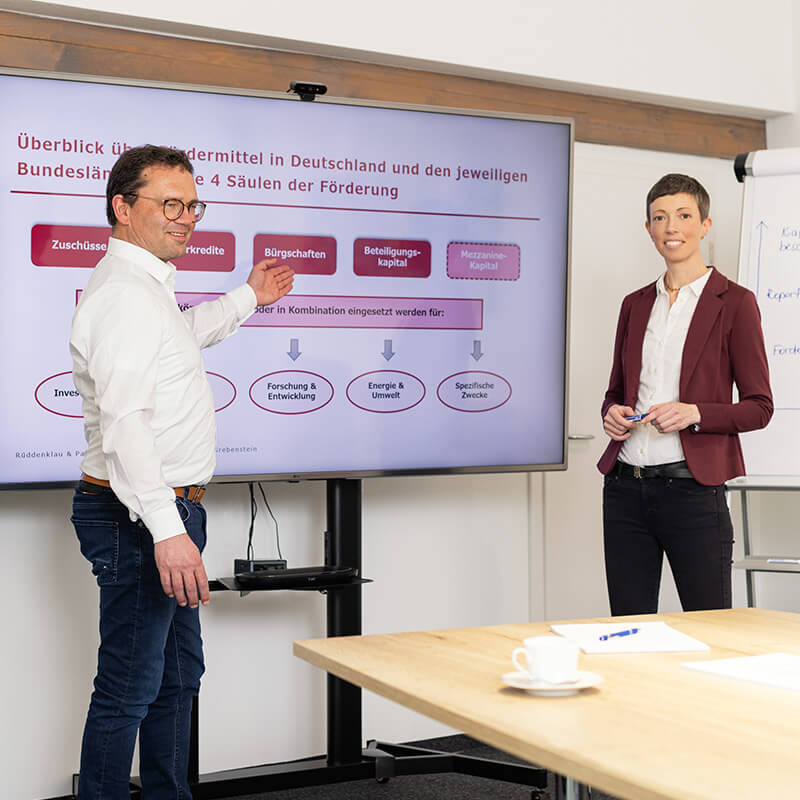 Auf dem Bild sieht man Marcus Kriegel und Dr. Monika Radloff in einem Konferenzraum. Zwischen ihnen sieht man eine Präsentation auf einem Bildschirm.